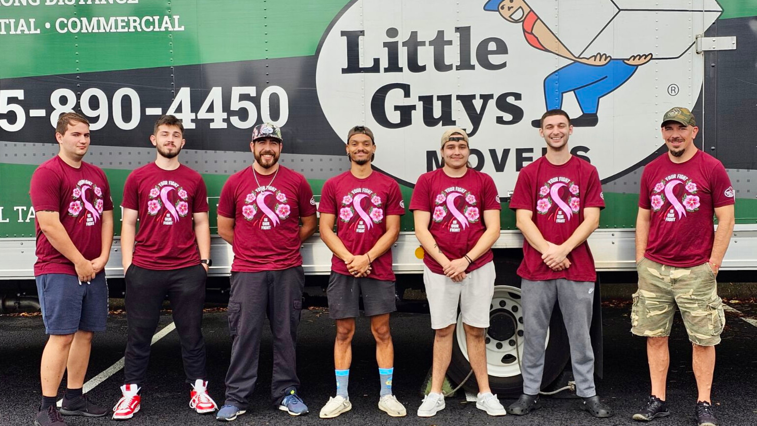 A crew of Little Guys Movers stand in front of a green moving truck wearing their pink Breast Cancer Awareness shirts.