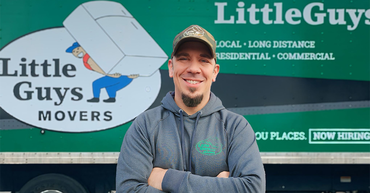 Little Guys Movers manager, Chris, wearing a grey hoodie and standing in front of a green moving truck.