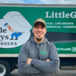 Little Guys Movers manager, Chris, wearing a grey hoodie and standing in front of a green moving truck.