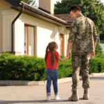 A man in military dress holds the hand of his daughter in front of a house.