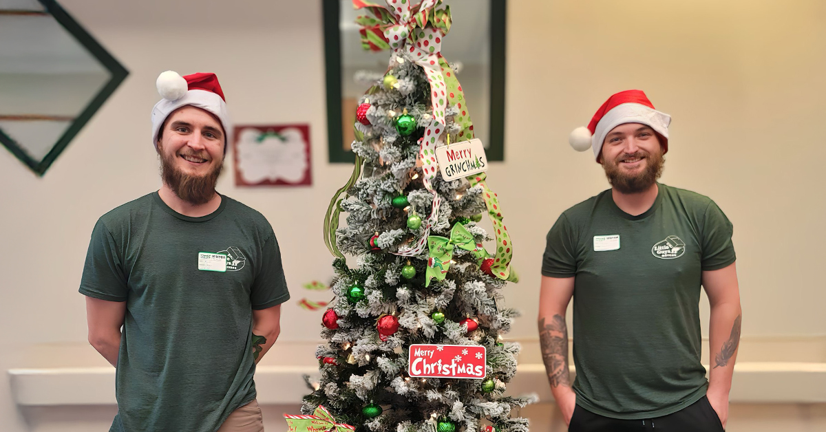 Two Little Guys Movers in green shirts stand beside the Grinch-themed Christmas tree they decorated