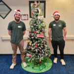 Two Little Guys Movers in green shirts stand beside the Grinch-themed Christmas tree they decorated